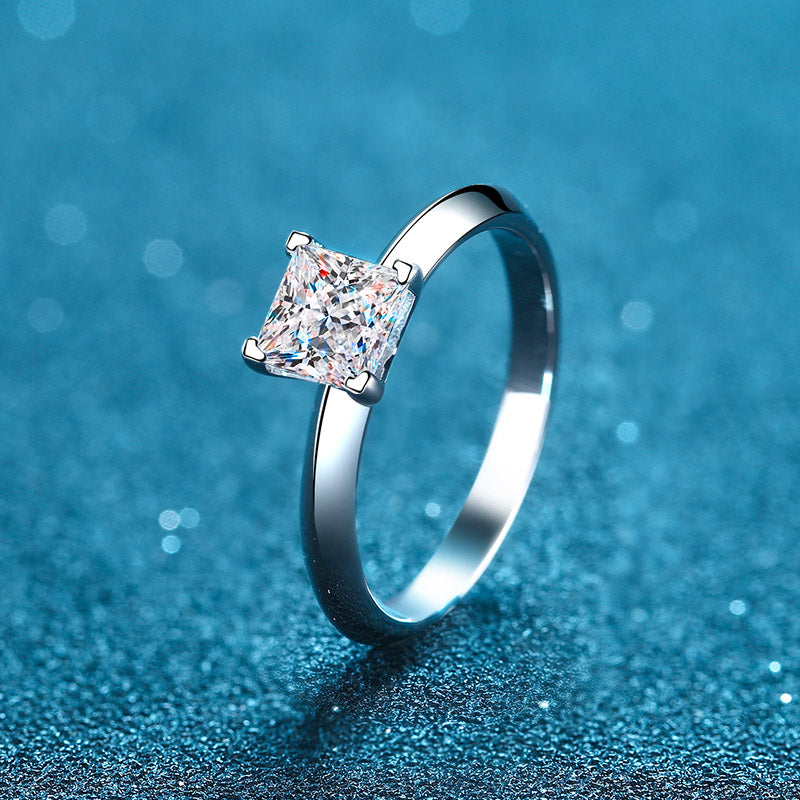 A princess cut solitaire moissanite diamond ring with a chevron pattern on a blue, sparkly background.