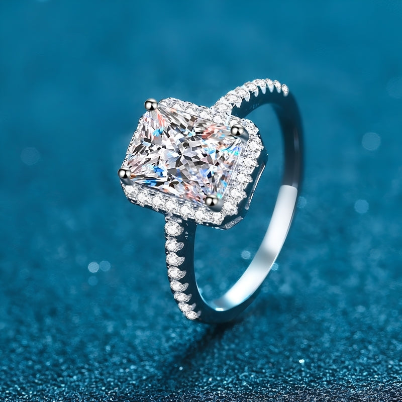 A silver-toned ring featuring a large square radiant-cut clear gemstone surrounded by a halo of smaller stones, with a pavé-set band, photographed on a textured blue background.